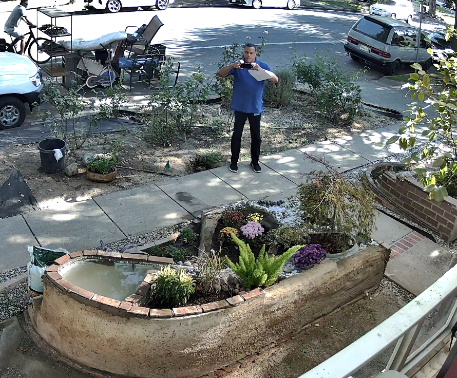 CCTV screenshot showing blue-shirt official standing on the sidewalk facing the house with phone held up.
